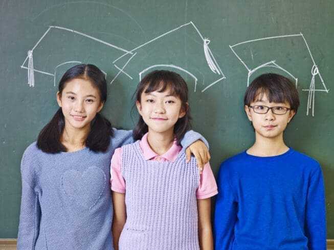 Who Invented School and Why: 5 Fascinating Traditions 8 three asian elementary school children standing in front of chalkboard underneath chalk-drawn doctoral caps.