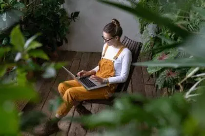 Focused millennial woman gardener in overalls watching educational webinar on laptop, writing blog or email, studying, remotely online work in her home garden surrounded by tropical plants. E-learning